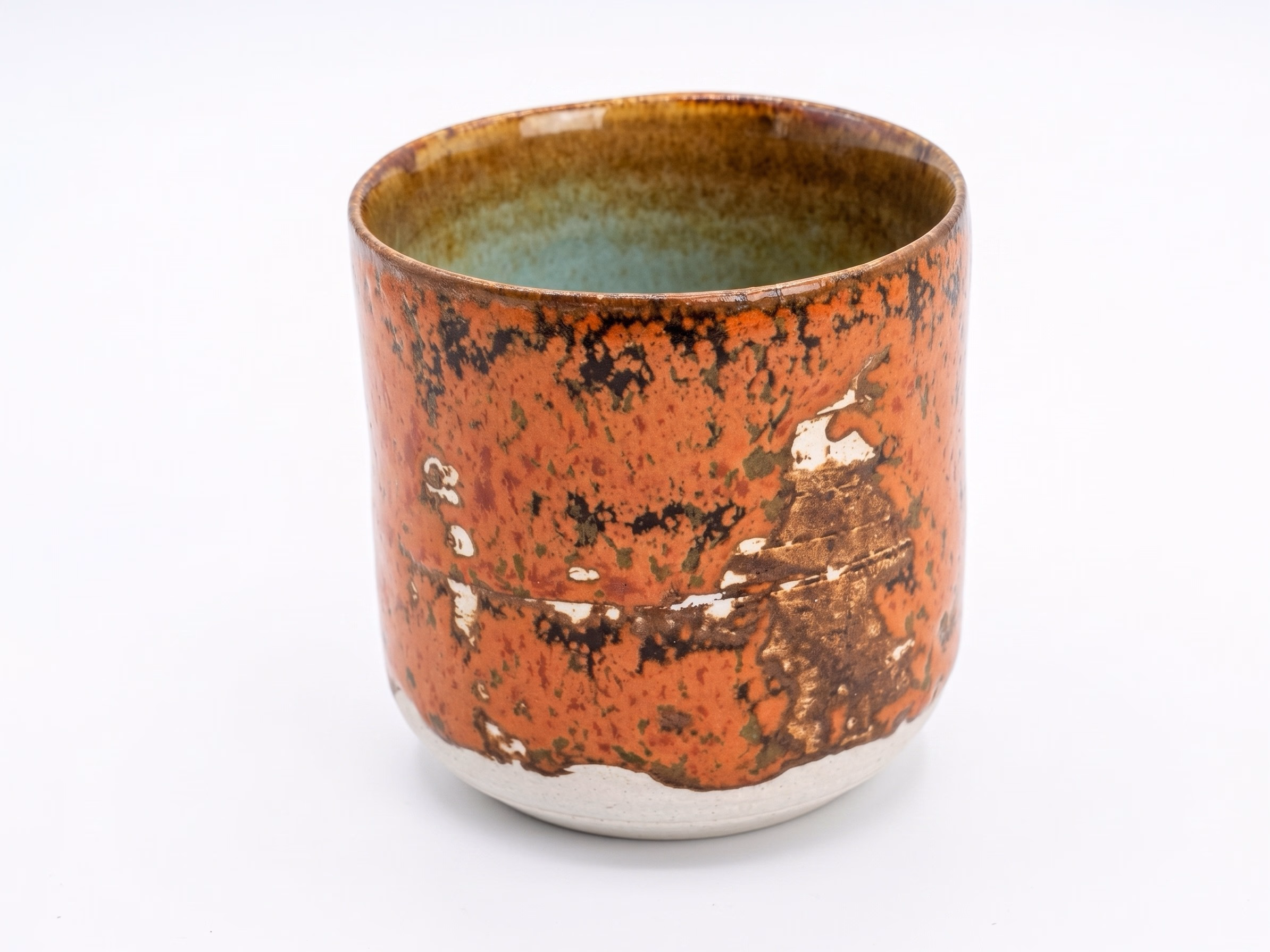 A cup in a heavily weathered iron-orange glaze with raw clay and pale revealed patches breaking through, soft green-grey interior.